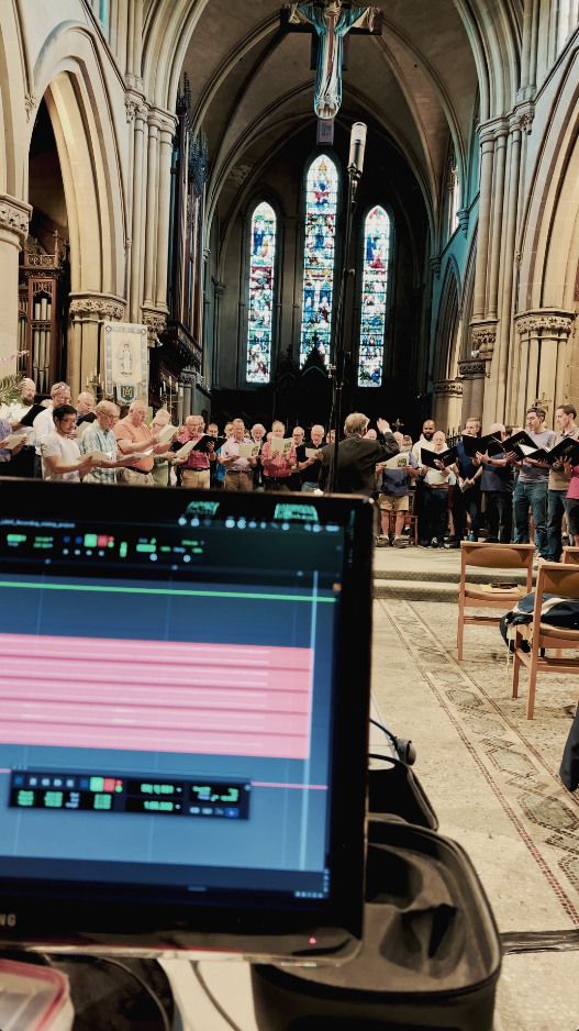 Leeds Male Voice Choir performing in a church, seen past a laptop running the multitrack recording session, with a microphone stand between the desk and the choir.
