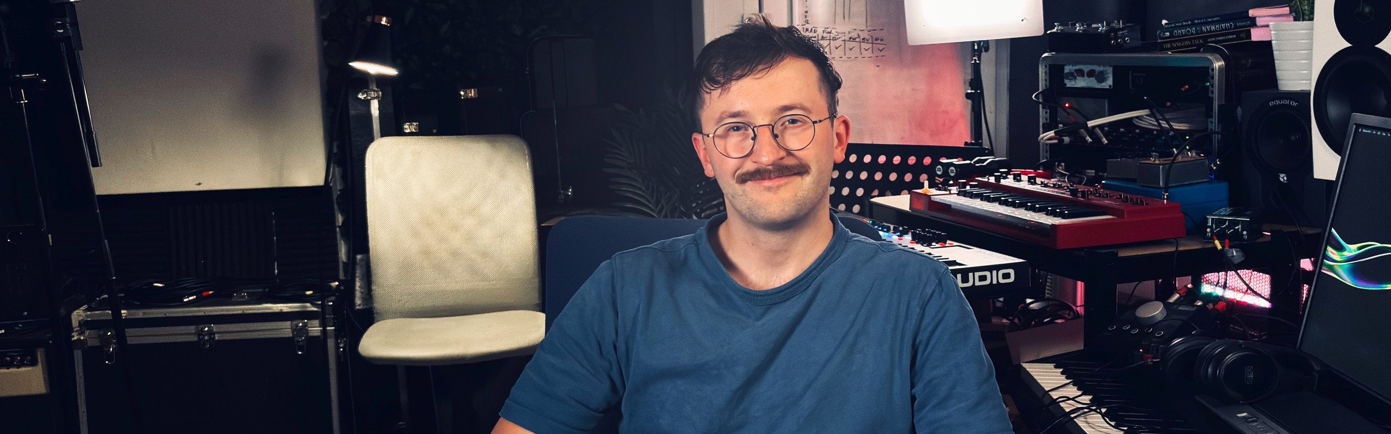 Max Greaves smiling at the camera in his studio, with keyboards, a red synth, an outboard rack and studio monitors behind him.