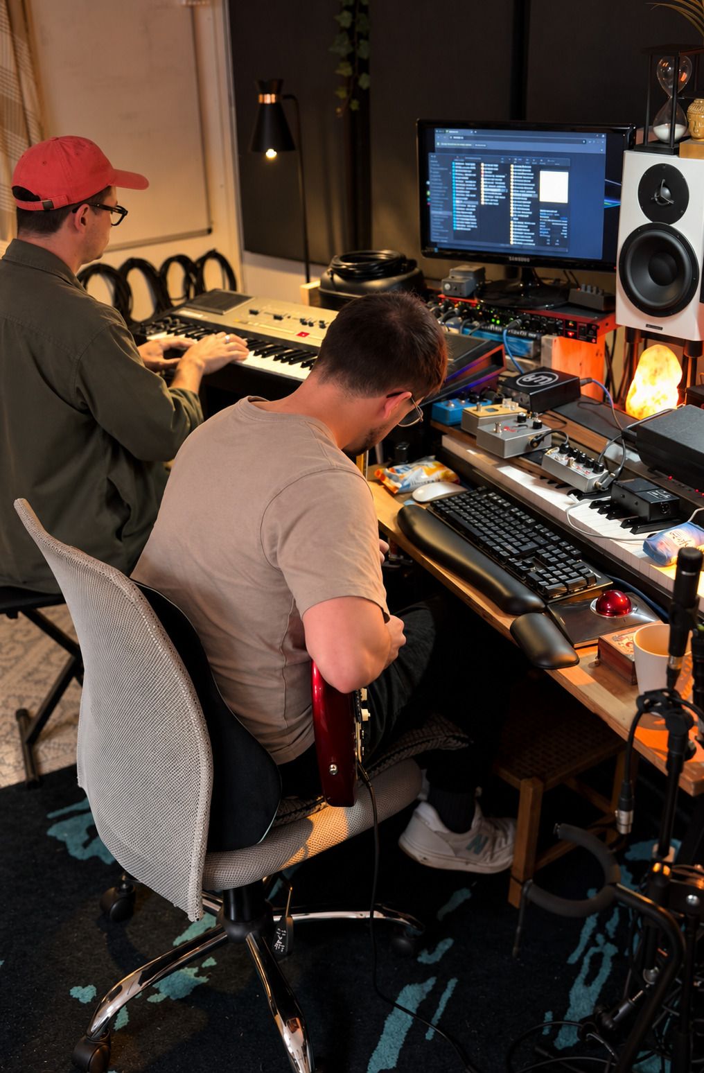 Two musicians at the studio desk, one playing a keyboard and one with a bass guitar, working through a part together.