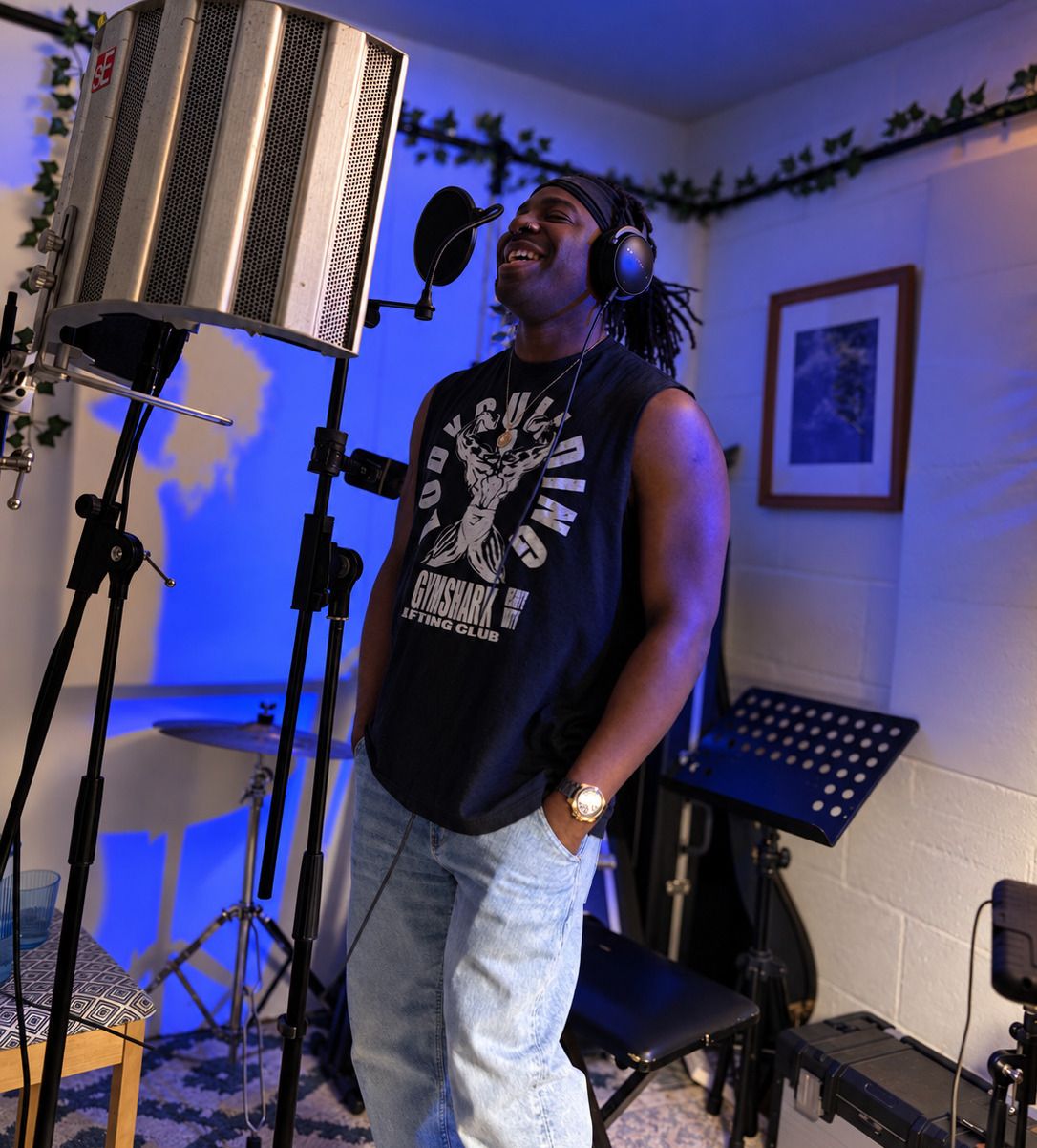 An artist in headphones laughing between takes in the vocal booth, lit in deep blue, with a reflection filter and pop shield in front of them.