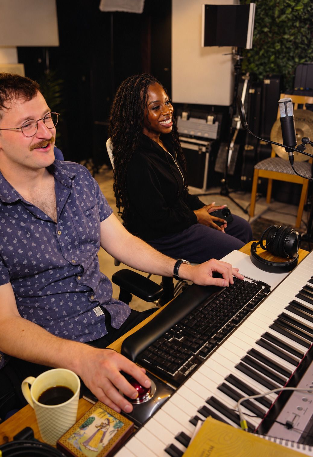 Max Greaves at a keyboard mid-laugh with artist IMAN beside him, a mug of coffee on the desk and cymbals and mic stands behind them.